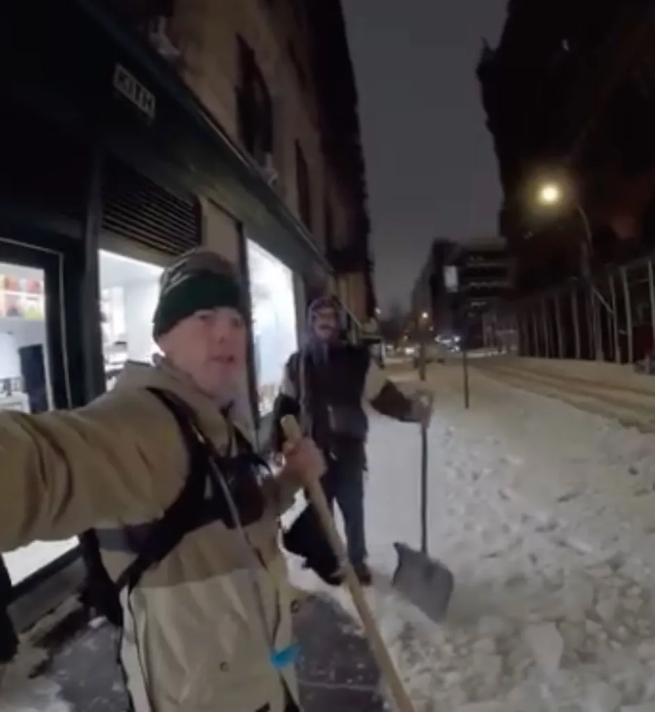 Two men holding shovels on a snow-covered street at night.